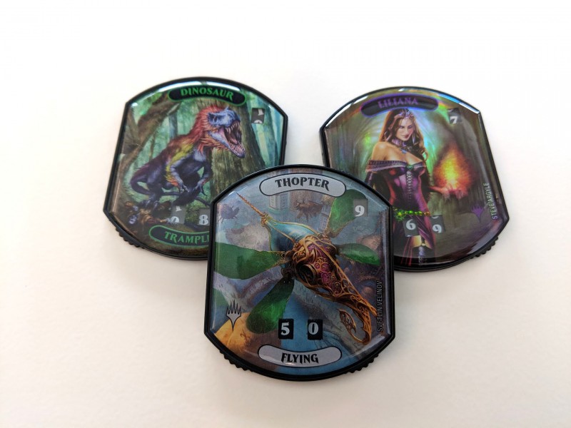 Three Magic: The Gathering token counters - one with a dinosaur on it, another with a thopter, and the third with Liliana.