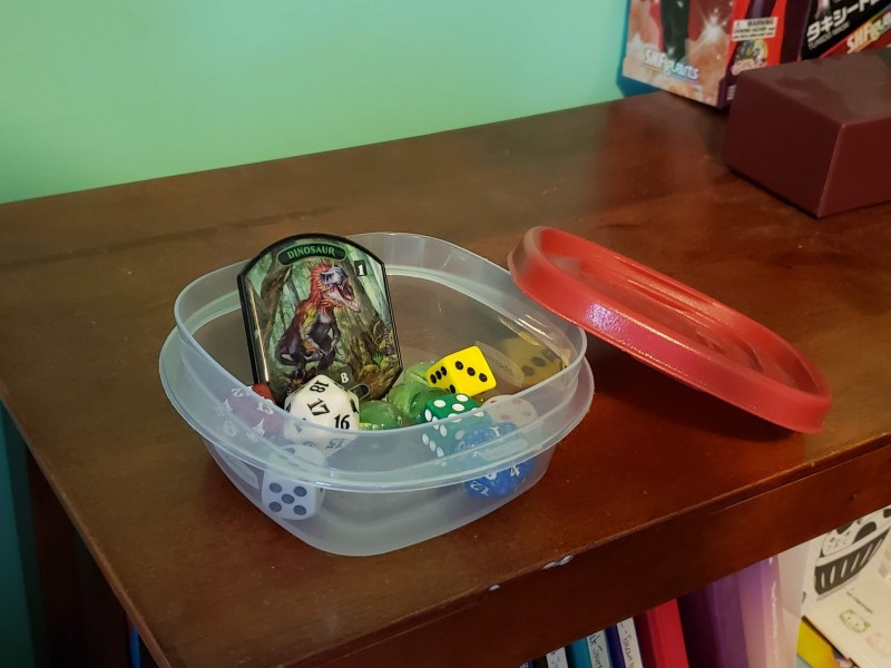 A tupperware container holding some small dice, a Magic: The Gathering health counter, and a dinosaur token counter.
