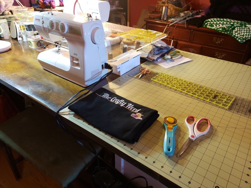 A table with a sewing machine, a Crafty Nerd t-shirt, and some fabric cutting tools laid out on top of it.