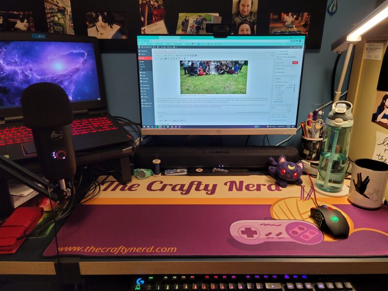 Photo of my desk with a large desk mat on it that has the text The Crafty Nerd across the top, and The Crafty Nerd's yarn ball and video game controller logo on the right side.